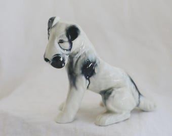 May include: A ceramic figurine of a seated dog with a glossy white finish and dark blue-grey mottled markings on its ears, face, and back. The dog is depicted in a sitting position with its head turned slightly to the left, featuring smooth curves.