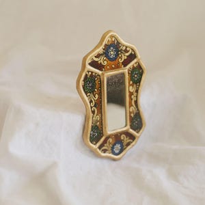 Puede incluir: Un peque&ntilde;o espejo decorativo con un marco dorado. El marco presenta un dise&ntilde;o ornamentado con motivos florales en tonos azules, verdes y marrones. El espejo tiene forma rectangular y est&aacute; sobre un fondo blanco.