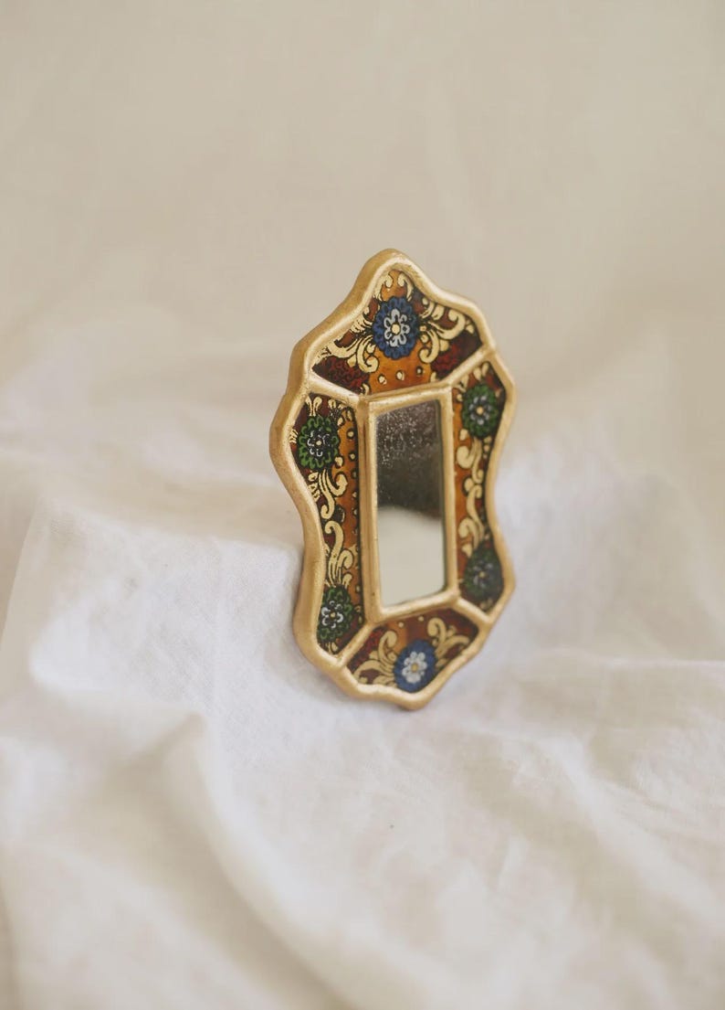 Puede incluir: Un peque&ntilde;o espejo decorativo con un marco dorado. El marco presenta un dise&ntilde;o ornamentado con motivos florales en tonos azules, verdes y marrones. El espejo tiene forma rectangular y est&aacute; sobre un fondo blanco.