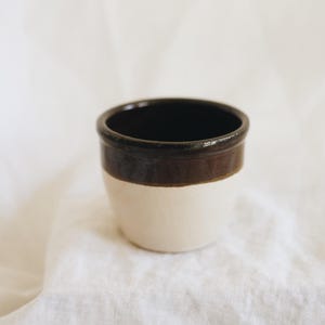 May include: A small, handcrafted ceramic cup with a two-tone design. The upper rim and a band around the middle are dark brown, while the lower portion is off-white. The interior is dark brown. The cup sits on a white, textured surface.