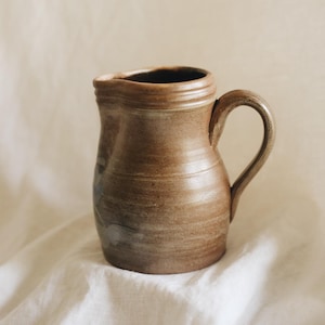 May include: A brown ceramic pitcher with a rounded body, a curved handle, and a wide pouring spout. The surface features subtle horizontal ridges and a smooth, earthy finish with faint blue-grey markings visible on the side of the vessel.