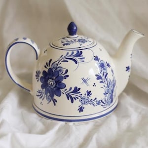 May include: A cream-colored ceramic teapot featuring hand-painted blue floral patterns and scrolling vines. The teapot includes a matching lid with a small blue knob, a curved handle, and a tapered spout, all decorated with blue line accents.