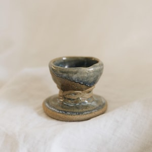 May include: A small, hand-thrown ceramic egg cup featuring a speckled, earthy brown base and a glossy, mottled blue-grey glaze on the upper rim. The piece has a wide, circular foot, a narrow neck with a textured band, and a flared, rounded bowl.