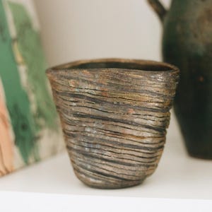 May include: A small, hand-thrown ceramic cup featuring a dark, earthy brown and charcoal gray finish. The exterior surface is defined by prominent, horizontal, ridged textures that wrap around the vessel, creating a layered, uneven appearance.