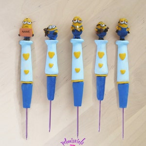 Puede incluir: Cinco ganchillos de crochet coloridos con un personaje de minion amarillo en la parte superior de cada gancho. Los ganchillos son azules, azul claro y amarillos. Los minions sostienen diferentes objetos, incluyendo un plátano, una guitarra y un osito de peluche. La palabra "NAME" está escrita en un pequeño ladrillo marrón que sostiene uno de los minions.
