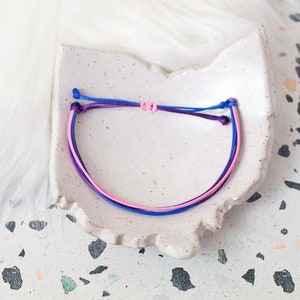 Puede incluir: Un brazalete de amistad hecho con cordón azul, rosa y morado. El brazalete está atado en un nudo y está sobre un plato de cerámica blanco.