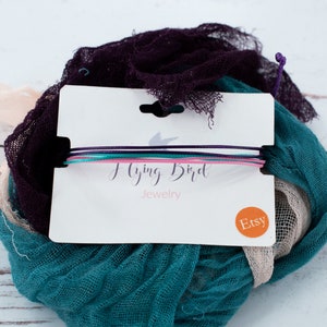 May include: A set of four colorful string bracelets on a white card with the text "Flying Bird Jewelry" and an Etsy sticker. The bracelets are purple, teal, pink, and white. The background includes teal, purple, and beige fabric.