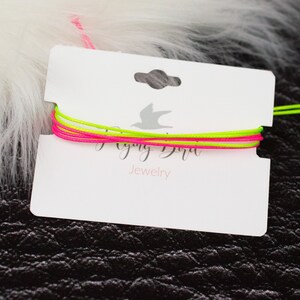 May include: A white card with the text "Flying Bird Jewelry" and a small bird logo. A neon green and pink string bracelet is displayed on the card.