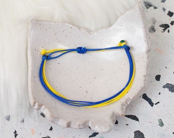 May include: A blue and yellow friendship bracelet with a knot on the side. The bracelet is on a white ceramic surface.