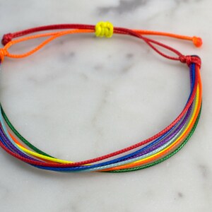 May include: A rainbow colored friendship bracelet made of thin cords tied together with a yellow knot.