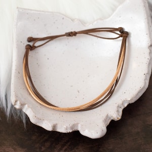 Puede incluir: Un brazalete de cuero marr&oacute;n con un acento de cuero marr&oacute;n claro. El brazalete est&aacute; atado con un nudo y se muestra en un plato de cer&aacute;mica blanca.
