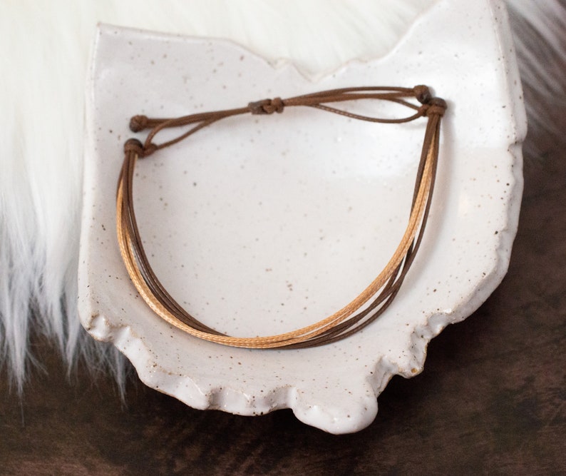 Puede incluir: Un brazalete de cuero marr&oacute;n con un acento de cuero marr&oacute;n claro. El brazalete est&aacute; atado con un nudo y se muestra en un plato de cer&aacute;mica blanca.