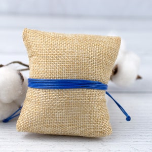 May include: A small, square burlap cushion tied with a blue ribbon. The cushion is sitting on a white wooden surface.