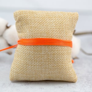 May include: A small, square, burlap pouch with a bright orange cord tied around it. The pouch is sitting on a white surface.