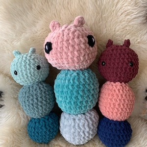 May include: Three crocheted stuffed animals shaped like caterpillars. The first caterpillar is light blue with a pink head, the second is pink with a blue head, and the third is maroon with a pink head. Each caterpillar has three body segments.