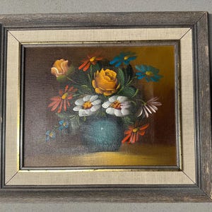May include: A framed floral still life painting. The artwork depicts a bouquet of colourful flowers in a teal vase. The flowers include yellow roses, white daisies, and red and blue accents. The painting is set against a dark background and framed in a rustic wooden frame.