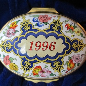 May include: An oval trinket box with a gold frame and a white enamel top. The top is decorated with a blue and yellow border, the red number "1996" and floral designs in pink, orange, and blue.