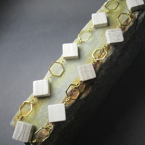 Peut inclure: Un collier en chaîne dorée avec des perles carrées blanches. Le collier est présenté sur une surface en pierre grise.