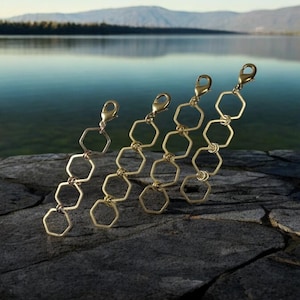 May include: Four gold-toned metal chains, each featuring a series of connected hexagonal links and a lobster claw clasp at the top. The chains are arranged in a row, displaying a uniform geometric design with open, six-sided metal shapes.