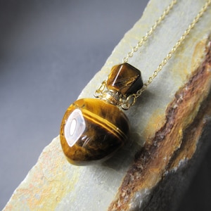 Puede incluir: Un collar con colgante de ojo de tigre en forma de corazón. El colgante es de color marrón cálido con detalles dorados y una cadena dorada. El collar se muestra sobre una superficie clara y texturizada.