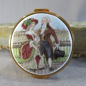 May include: A round, gold-toned compact featuring a painted scene of a couple in 18th-century attire. The woman is wearing a red and green dress and a red hat. The man is wearing a blue and red suit and a white wig. They are standing in front of a large building with a green lawn.