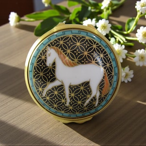 May include: A round gold-toned compact mirror featuring a white horse with a tan mane and tail centered on a black background with a gold geometric pattern. The edge is decorated with a teal border featuring a repeating Greek key design.