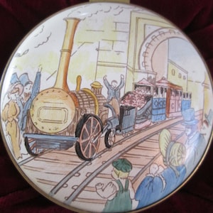May include: A vintage-style round tin box with a colorful illustration of a steam train pulling carriages. The train is on a track with people watching from the side. The tin box has a gold rim.