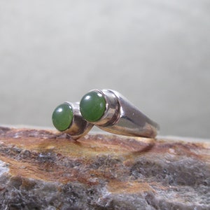 Puede incluir: Un anillo de estilo bypass hecho de metal plateado pulido, con dos piedras cabujón redondas, lisas y de color verde oscuro engastadas en biseles individuales. El anillo tiene una banda elegante y reflectante que se estrecha hacia atrás.