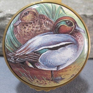 May include: A round brass-rimmed decorative object featuring a detailed illustration of two ducks. One duck has brown mottled feathers, while the other displays a chestnut head with a green eye patch and a light blue wing patch, set against green grass.