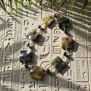 Puede incluir: Un collar con piedras de jade cuadradas multicolores, unidas por una cadena dorada. Pequeñas cuentas negras acentúan el diseño. Se exhibe sobre una superficie texturizada blanquecina con patrones jeroglíficos.