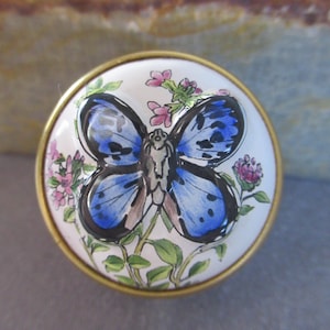 Puede incluir: Un pomo de cerámica blanca con un borde dorado presenta una mariposa azul con marcas negras y acentos blancos, rodeada de flores rosas y verdes.