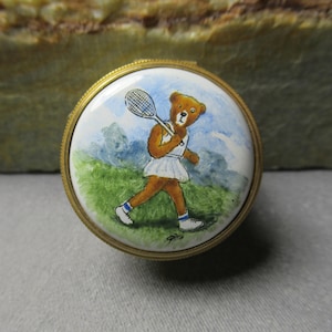 Op de afbeelding: Een ronde, witte porseleinen sieradenbox met een gouden rand. Het deksel heeft een handgeschilderd beeld van een bruine teddybeer die een witte tennisjurk draagt en een tennisracket vasthoudt. De beer loopt op een groen grasveld met een blauwe lucht en witte wolken op de achtergrond.