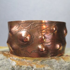 May include: A copper cuff bracelet with an embossed floral design and raised, rounded accents. The bracelet has a warm, reddish-brown hue and a textured surface, showcasing the intricate details of the metalwork.