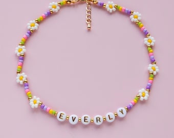 May include: A white beaded necklace with colorful beads and white daisy charms. The necklace has the name "EVERLY" spelled out in gold letters on white beads.