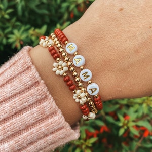 May include: A set of three beaded bracelets featuring small red and metallic gold beads, white flower-shaped beads, and white round beads with gold lettering that spells out MAMA. The bracelets are worn together on a wrist with a pink sweater sleeve.