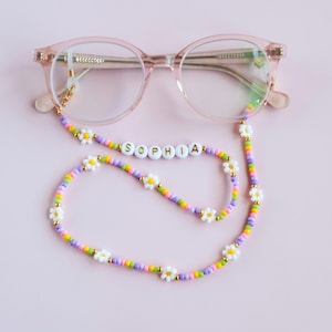 May include: A pink and white beaded glasses chain with white daisy beads and gold letters spelling "SOPHIA". The chain is attached to a pair of pink glasses.