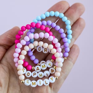 May include: A collection of four elastic bracelets made of matte spherical beads in light blue, purple, hot pink, and pale pink. Some bracelets feature white circular beads with gold-colored letters spelling out names like EMILY, SOPHIA, and AMY.