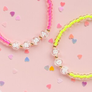 Könnte beinhalten: Zwei Armbänder mit Perlen, auf denen die Buchstaben "MAMA" und "BRIDE" stehen. Ein Armband ist neonrosa, das andere neongelb. Goldfarbene Perlen trennen die Buchstabenperlen. Herzförmiges Konfetti ist auf einem rosa Hintergrund verstreut.