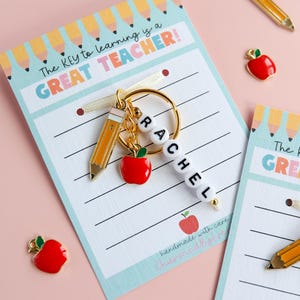 Puede incluir: Un llavero con un dije de lápiz y manzana, deletreando "RACHEL" con cuentas de letras blancas. El llavero cuelga de una tarjeta que dice "The KEY to learning is a GREAT TEACHER!" y presenta un diseño de papel rayado.