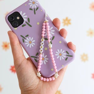 Può includere: Una custodia per telefono viola con un motivo di margherite bianche e un cinturino per telefono con perline rosa con accenti dorati e perline a forma di stella bianca. Il cinturino ha la parola "MAMA" scritta in lettere dorate.