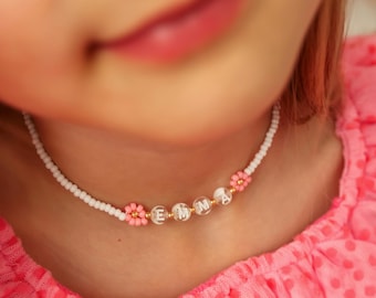 May include: A white beaded necklace with the name "EMMA" spelled out in clear letter beads. Pink flower accents are interspersed throughout the necklace. The necklace is worn by a person wearing a pink top.