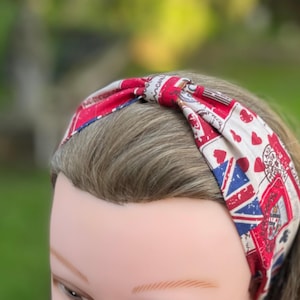 May include: A fabric headband featuring a red, white, and blue pattern with Union Jack flags, scattered red hearts, and vintage-style postage stamp graphics. The headband has a knotted center detail and is worn on a mannequin head.