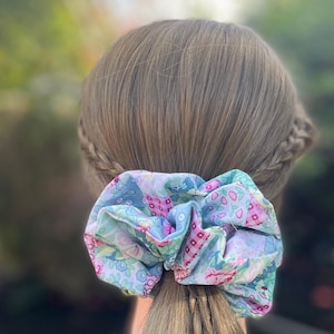 May include: A large scrunchie in a ponytail, featuring a patchwork design with pink, blue, and green floral patterns. The hair is light brown and styled in a braid.