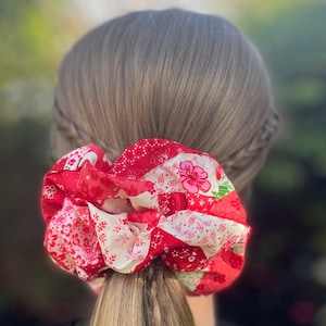 May include: A large, red scrunchie with a patchwork design, featuring floral and geometric patterns in shades of red and white. The scrunchie is made of fabric and is worn in a ponytail.