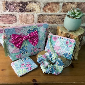 May include: A set of fabric accessories featuring a patchwork floral pattern in shades of blue, pink, and green. The set includes a large zippered pouch with a pink polka-dot bow, a smaller zippered pouch, a square fabric pad, and a hair scrunchie.