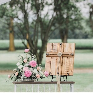 May include: A wooden sign with a rope cross design, personalized with the names "Blake & Ashley" and the date "August 17, 2023". The sign is displayed on a black metal stand and decorated with a floral arrangement of pink and white flowers.