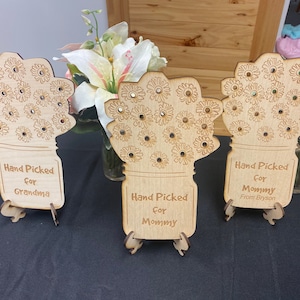 May include: Three wooden flower bouquet crafts with a mason jar design. Each bouquet has a different message: "Hand Picked for Grandma", "Hand Picked for Mommy", and "Hand Picked for Mommy from Bryson".