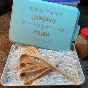 May include: A light blue cake pan with the text "The Secret Ingredient Grandma's Kitchen Is Always Love" on the lid. The pan is open and contains a set of four wooden measuring spoons with the text "My favorite treats come from Grandma's Kitchen" and "Measured with Love" on them. The spoons are nestled in white shredded paper.