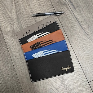 May include: Five leather checkbook covers with different designs. The top cover is gray with the text "Be Still" in black. The second cover is brown with a white and black American flag design. The third cover is blue with a white American flag design. The fourth cover is black with a white American flag design. The bottom cover is black with the name "Angela" in gold lettering.