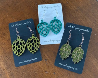 May include: Three pairs of green and gold hop-shaped earrings on black cards with the text "C&A Engraving and Gifts" and the website address "www.candaengraving.com".